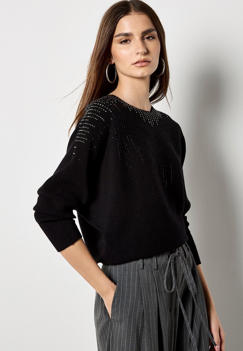 Schwarzer gestrickter Pullover mit einem strukturierten Muster; hat einen lockeren Schnitt und weite Ärmel. Kombiniert mit grauen gestreiften hochtaillierten Hosen.