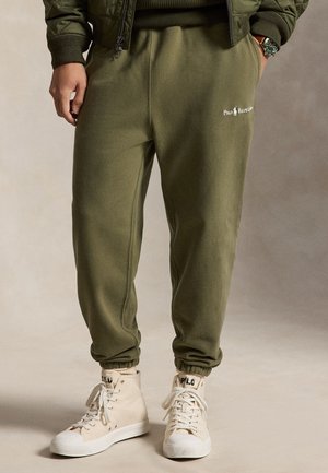 Άνδρας που φορά χακί ελαστικό παντελόνι Polo Ralph Lauren και μπεζ ψηλά αθλητικά παπούτσια με κορδόνια, στέκεται μπροστά σε ουδέτερο φόντο.