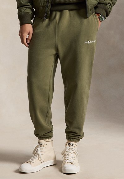 Άνδρας που φορά χακί ελαστικό παντελόνι Polo Ralph Lauren και μπεζ ψηλά αθλητικά παπούτσια με κορδόνια, στέκεται μπροστά σε ουδέτερο φόντο.