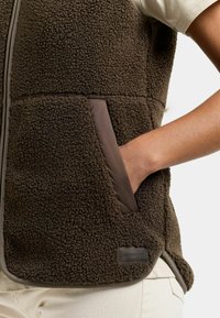 Hand in de zak van een donkerbruine, gestructureerde fleece-vest gedragen over een licht overhemd en crème broek, met details van de stof en de zak van de vest zichtbaar.