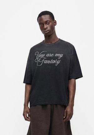 Fiatal férfi laza fekete pólóban, amin a "You are my Fantasy" szöveg dőlt, fehér betűkkel van a mellkasán.