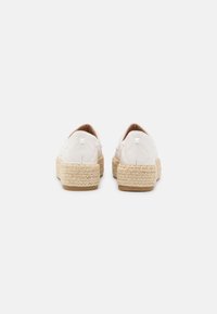Baskets blanches à enfiler avec une texture matelassée et des clous carrés à l'arrière, présentant des semelles en jute tressé, vues de l'arrière sur fond blanc.