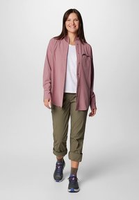 Lys rosa skjorte med knapper over en hvit t-skjorte, kombinert med opprullede olivengrønne bukser og mørke joggesko med lilla detaljer. Har en brystlomme.