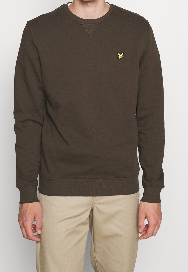 Homme portant un sweat-shirt vert olive foncé à col rond avec un petit logo d'oiseau jaune sur la poitrine et un pantalon beige, debout devant un fond blanc.