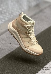 Beige Wanderschuh mit einer strukturierten Oberseite und Netzpaneelen, Schnürverschluss und Gummisohle mit Profil, schwebend vor einem grauen Hintergrund.