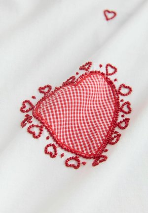Balts audums ar sarkanas rūtainas sirds dizainu, ko ieskauj smalkas sarkanas izšuvumi ar mazām sirsniņām gar malām.