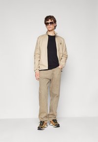 Giacca leggera beige con colletto alto, maglietta nera a girocollo, pantaloni beige chiaro e sneakers nere/gialle. Vengono indossati occhiali da sole.