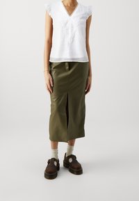 Blusa bianca con pizzi, maniche corte a volant, abbinata a una gonna in pelle verde oliva fitted con uno spacco frontale. Scarpe marroni con cinturini.