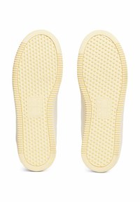 Semelles de chaussures en caoutchouc de couleur claire avec un motif texturé en triangles et des bords arrondis. Marquage présent au centre de chaque semelle.