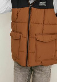 Brązowa kamizelka puchowa z czarną górną częścią, z zamkiem błyskawicznym z przodu i dwiema dużymi kieszeniami z przodu. Materiał wydaje się pikowany.