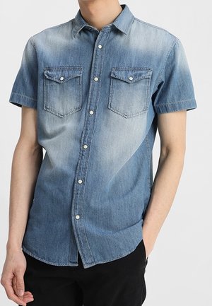 Mann trägt ein kurzärmeliges, knöpfbares Jeanshemd mit zwei Brusttaschen und schwarze Hosen, steht mit einer Hand in der Tasche.