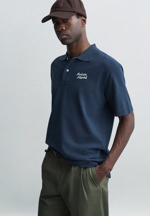 Marineblaues Poloshirt aus strukturiertem Stoff mit einem Kragen mit drei Knöpfen, versehen mit einem weißen gestickten Logo. Braune Kappe mit gebogenem Schirm.