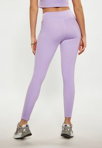 Lila leggings magas derékkal, rugalmas anyagból készült, sima textúrával és testhezálló fazonnal, szürke edzőcipőkkel párosítva.