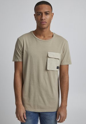 Beige cotton t-shirt with a round neckline, short sleeves, and a front pocket. Features a tag on the pocket. Worn with blue denim jeans.