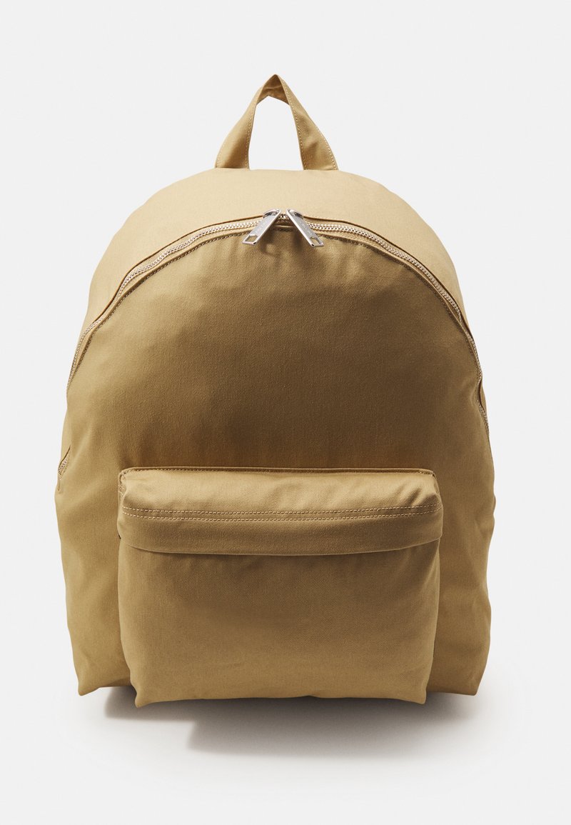 Beige stoffen rugzak met een ronde vorm, bovenhandgreep, dubbele ritsluiting en een voorvak. Gladde textuur met minimale stikdetails.