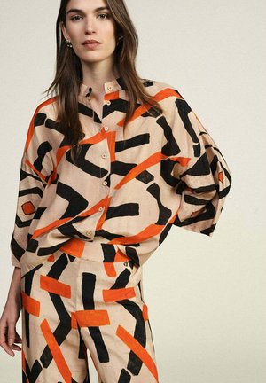 Donna che indossa una camicia beige a taglio ampio e pantaloni coordinati con audaci motivi geometrici astratti neri e arancioni, in piedi con la mano sul fianco.