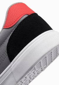 Šedé semišové tenisky s černými a korálovými akcenty, texturovaná tkanina, gumová podrážka a vzorovaná podrážka pro lepší přilnavost. Detailní design paty je viditelný.