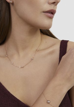 Close-up of woman wearing a delicate gold necklace and matching bracelet with small round gemstones, dressed in a burgundy sleeveless top.