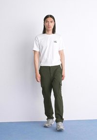 Hombre con cabello largo y negro, vistiendo una camiseta blanca, pantalones cargo verde oliva y zapatillas grises, de pie sobre un suelo azul contra una pared blanca.