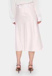 Jupe midi évasée en satin rose doux, présentant une texture lisse et une taille. Portée avec des sandales à talons hauts à lanières.