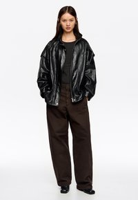 Mujer con cabello oscuro y largo, vistiendo una chaqueta de imitación cuero negra de gran tamaño, una blusa negra de canalé, pantalones marrones anchos y zapatos negros sobre fondo blanco.