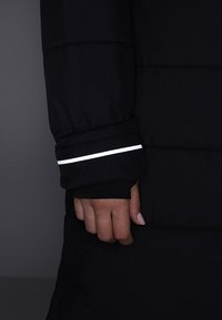 Mano con manga de chaqueta de plumón negra con una franja blanca reflectante cerca de la muñeca contra un fondo oscuro.