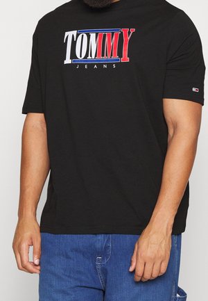 Hombre con una camiseta negra con el logo de "TOMMY JEANS" en blanco, azul y rojo, combinado con vaqueros de mezclilla azul.