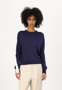 Felpa blu scuro con maniche lunghe, dotata di polsini e orlo a coste, e un sottile logo in rilievo sulla parte anteriore. Abbinata a pantaloni beige chiaro.