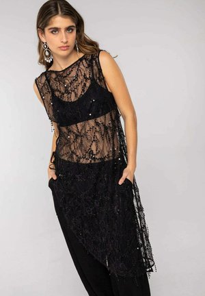 Joven con una blusa negra sin mangas de encaje transparente con lentejuelas sobre un sujetador negro y pantalones negros, de pie con las manos en los bolsillos.