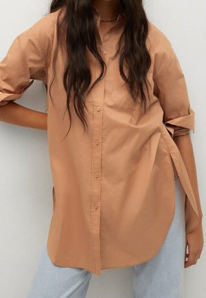 Kobieta ubrana w luźną, beżową koszulę na guziki z długimi rękawami podwiniętymi do łokci oraz jasnoniebieskie dżinsy, stojąca na jednolitym tle.