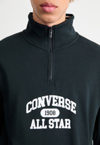 Zwarte fleece kwart-ritssweater met een hoge kraag. Bevat een witte "CONVERSE ALL STAR" logo en een ritsopening bij de nek.