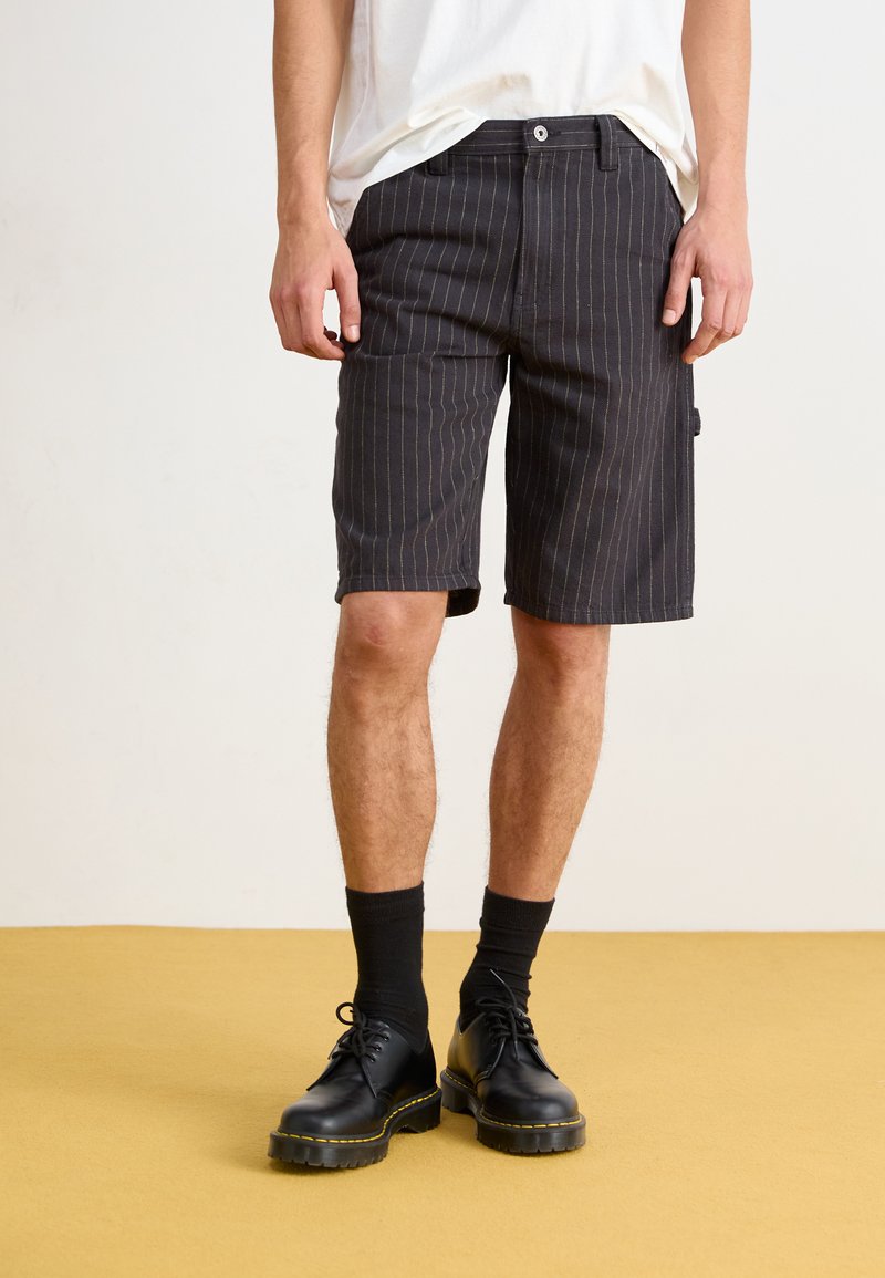 Svarte stripete shorts laget av teksturert stoff, med sidelommer, knappelukking og en avslappet passform, paret med svarte sko og sokker til ankelen.