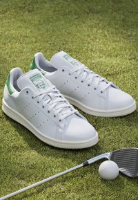 Bílé tenisky s zelenými akcenty, hladký kožený svršek, kulatá špička, perforované boky a plochá podešev. Obsahuje branding Adidas na jazyku.