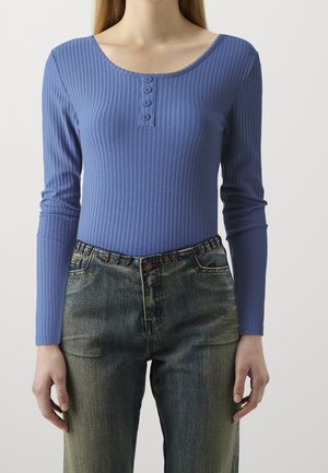 Kobieta ubrana w dopasowaną, niebieską, prążkowaną bluzkę z długim rękawem, zapinaną na cztery guziki, oraz wyblakłe, wysokowalne dżinsy z metalowymi oczkami.