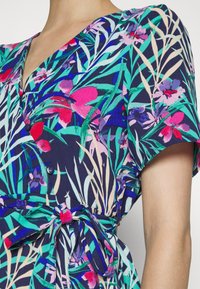Vestido floral com base azul-marinho, apresentando padrões de plantas coloridos, mangas curtas, decote em V e detalhe de laço na cintura com botões.