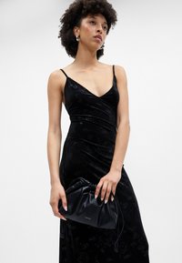 Melnas zīda kleitas ar V veida kakla izgriezumu, šaurām siksniņām un ziedu rakstu. Modelis tur melnu ādas somiņu ar savilkumu detaļām.