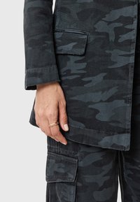 Giacca in tessuto mimetico scuro con tasca, caratterizzata da una silhouette sartoriale. È visibile una mano adornata da un semplice anello d'oro.