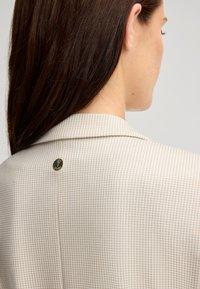 Beżowa marynarka w wzór houndstooth z wyciętym kołnierzem, z małym złotym logo z tyłu. Gładka tekstura z wyraźnymi liniami.