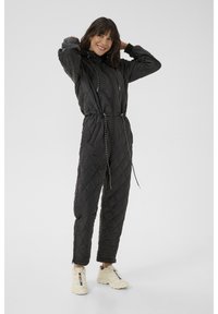 Schwarzer gesteppter Overall mit Reißverschluss vorne, verstellbarem Kordelzug in der Taille und elastischen Bündchen. Kombiniert mit hellen Sportschuhen.