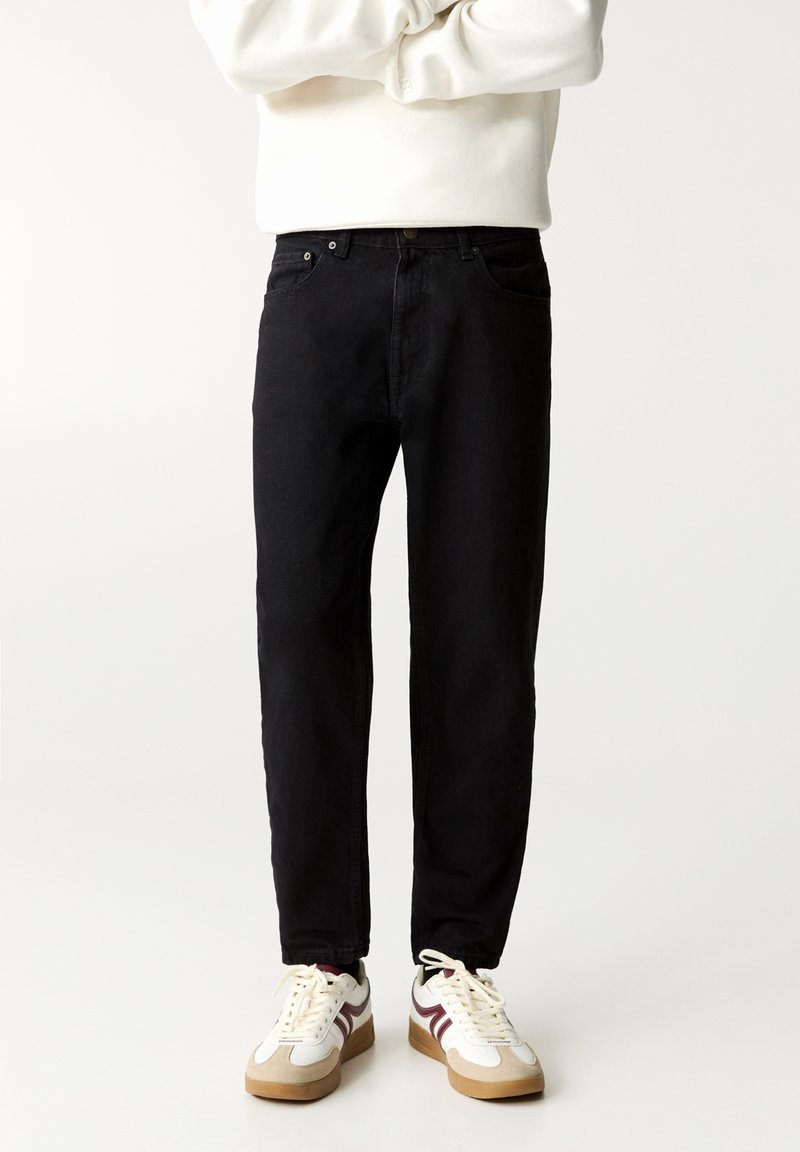 Personne portant un pantalon noir, des baskets blanches avec des bandes marron et un haut blanc à manches longues, debout les bras croisés.