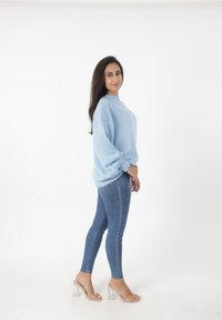 Suéter de punto azul claro con un cuello alto y mangas anchas, combinado con jeans skinny azules y sandalias de tacón transparente. Modelo de pie de perfil.