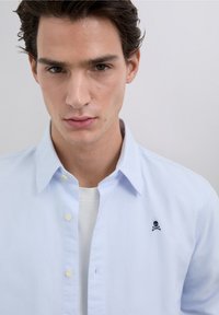 Světle modrá košile na knoflíky s jemnou texturou a malým černým logem lebky a zkřížených kostí na hrudi. Nošená přes bílé tričko.