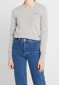 Hellgrauer Pullover mit langen Ärmeln, Rundhalsausschnitt und gerippten Bündchen. Auf der Brust befindet sich ein kleines rotes Logo. Kombiniert mit blauen, hochgeschnittenen Jeans.