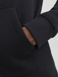 Black sweatshirt with a soft texture, featuring a kangaroo pocket and ribbed cuffs. Close-up view of the hand resting in the pocket.