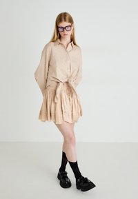 Chemise boutonnée à carreaux beiges nouée à la taille, associée à une jupe beige à couches et des chaussures noires imposantes, complétées par des chaussettes montantes.