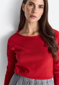 Vrouw met lang bruin haar, gekleed in een rode top met lange mouwen en een grijze rok, staande tegen een effen witte achtergrond.