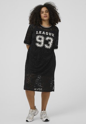 Kobieta o kręconych włosach, ubrana w czarną koronkową sukienkę z napisem "LEAGUE 93" i białe tenisówki, stojąca na jednolitym jasnym tle.