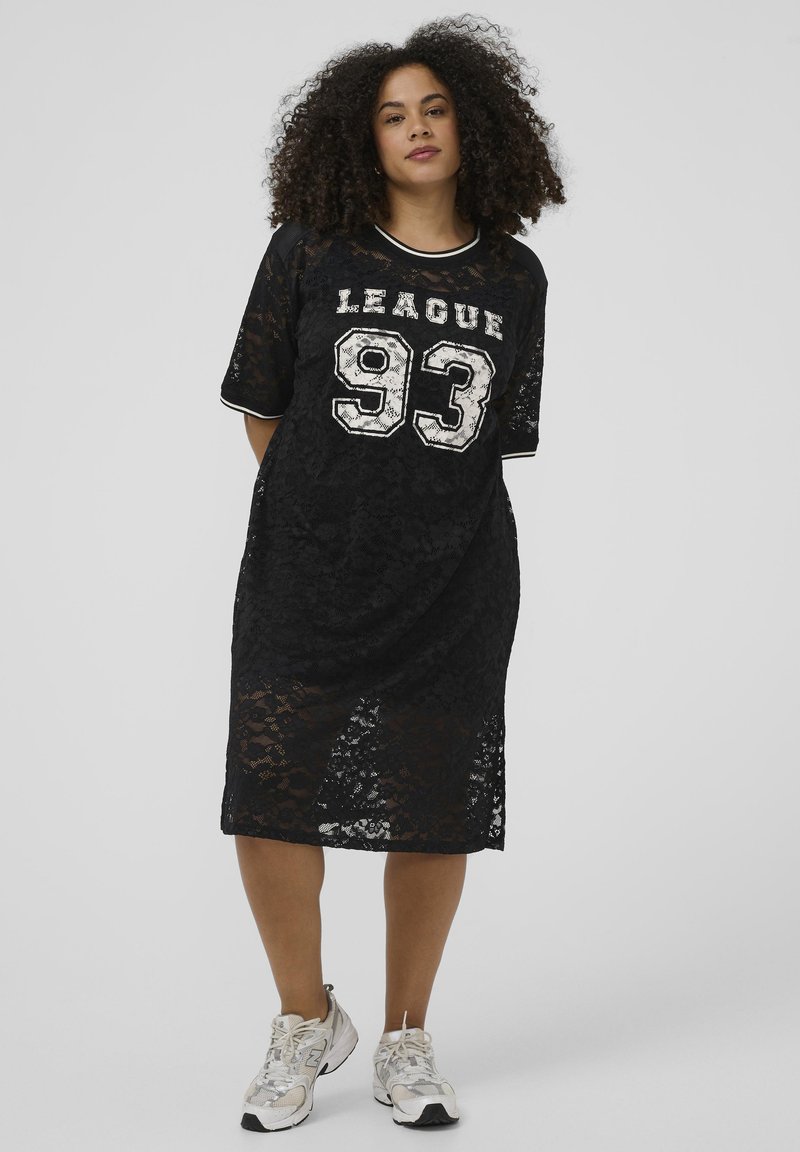 Femme aux cheveux bouclés portant une robe en dentelle noire avec le texte « LEAGUE 93 » et des baskets blanches, se tenant contre un fond clair uni.