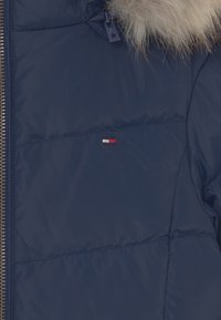 Mörkblå pufferjacka med huva i päls. Har en synlig dragkedja och en liten logotypspatch i rött, vitt och blått på bröstet.