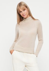 Pull-over beige à col roulé en tricot léger. Il présente des manches longues, des poignets côtelés et une silhouette ajustée. Texture lisse, design minimal.